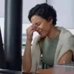 Mulher profissional no escritório com a mão no rosto, cobrindo os olhos com expressão de dor de cabeça, exaustão e cansaço em frente ao computador.