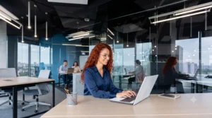 Mulher jovem com cabelos ruivos cacheados e óculos, sorrindo e digitando em um notebook em um escritório moderno de plano aberto. Ela parece estar consultando informações online sobre crédito do trabalho, consultoria de carreira ou gerenciamento financeiro.